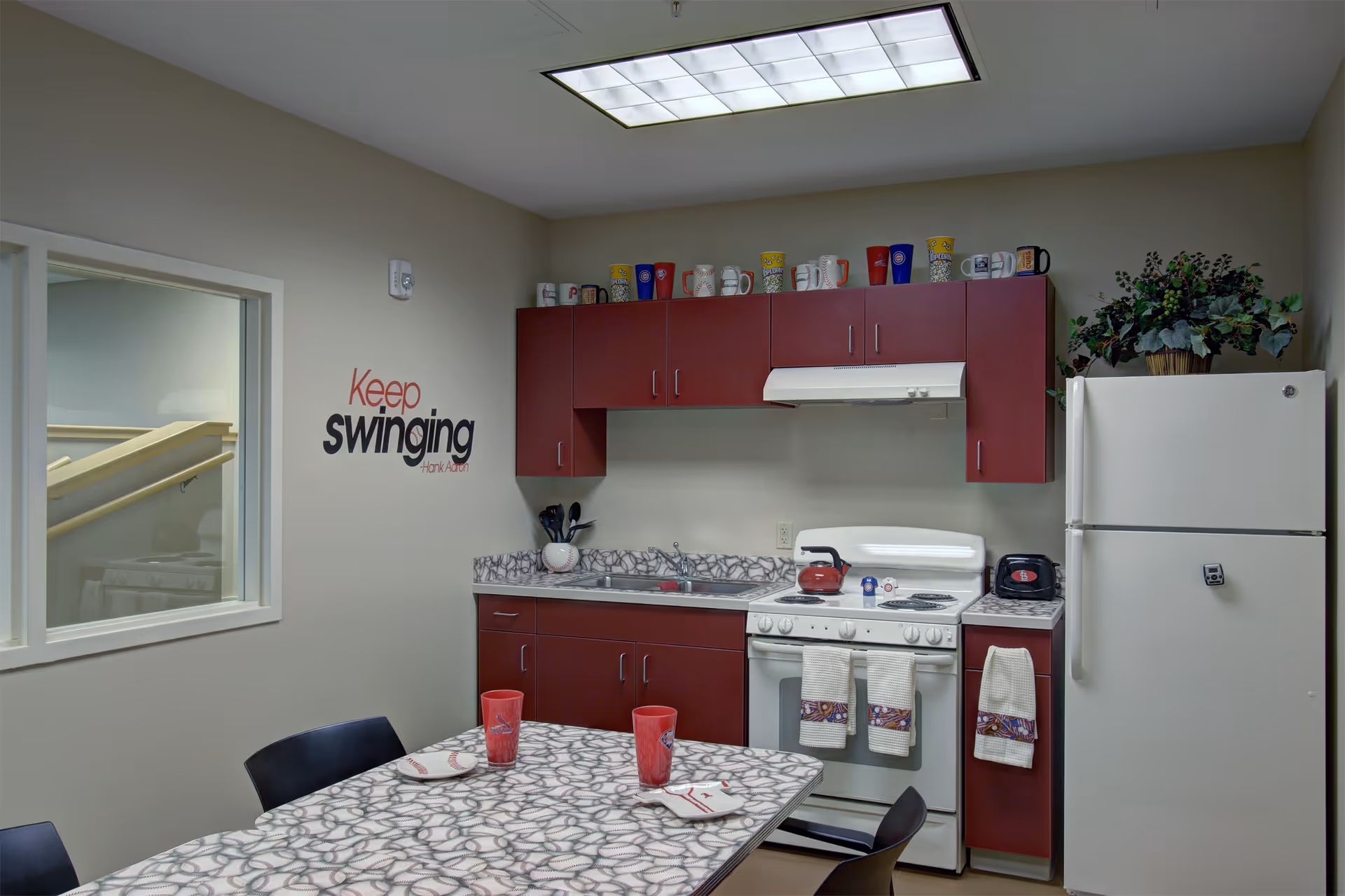 A small kitchen area with maroon cabinets, a white stove with a red kettle, a white refrigerator, and a countertop with a sink. On the wall, there is a quote that says 'Keep swinging' by Hank Aaron. A table with a patterned surface has two red cups and two plates with utensils. There are several mugs displayed on top of the cabinets and a potted plant on the refrigerator.