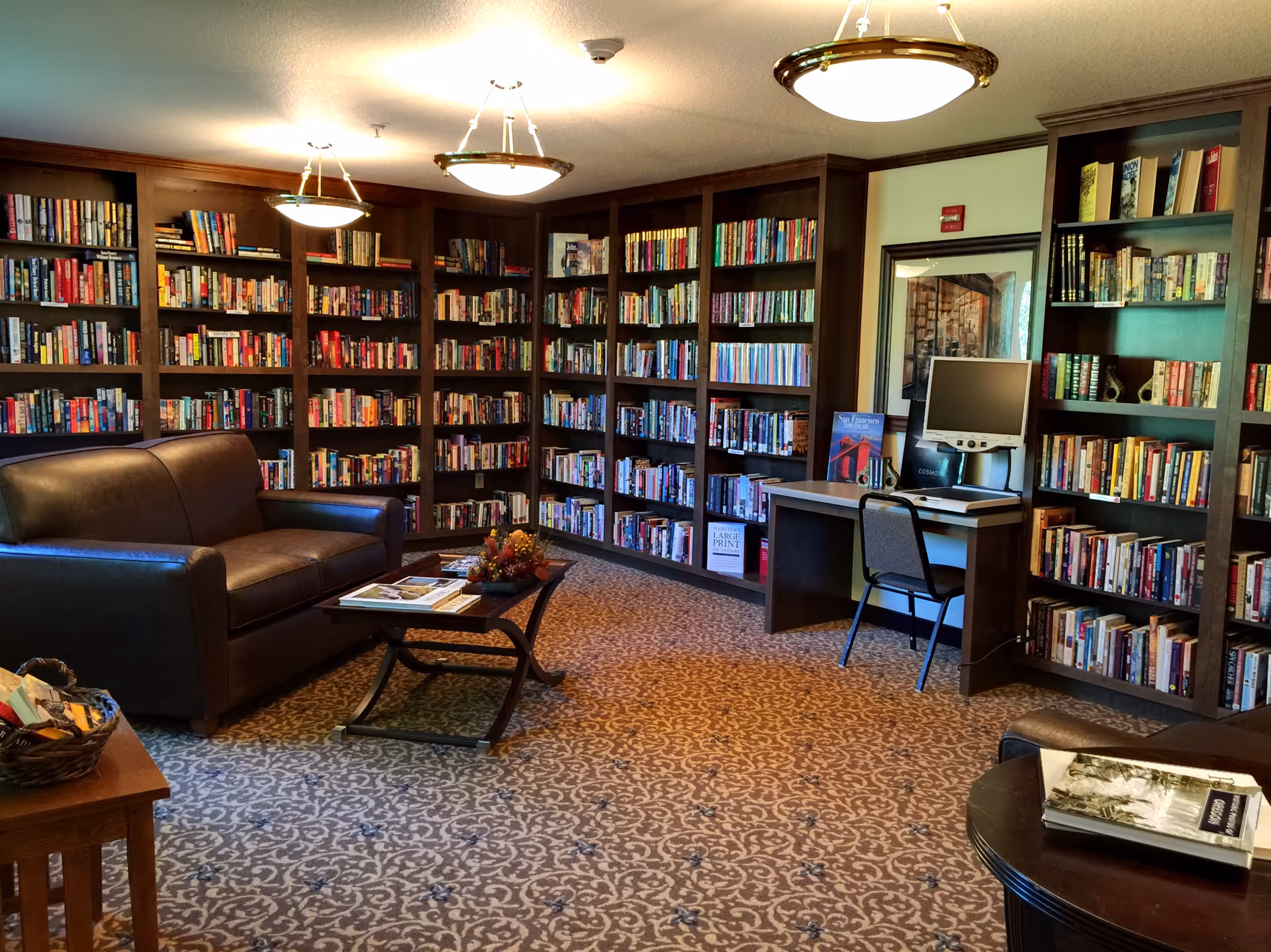 A cozy library room with dark wooden bookshelves filled with books lining the walls. There is a brown leather couch and a coffee table with magazines and a decorative centerpiece. A computer desk with a monitor and chair is positioned against one wall. The room is carpeted with a patterned design and lit by ceiling lights.