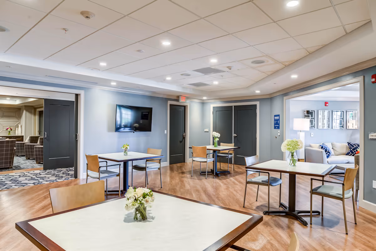 A bright and spacious common area in a senior living facility with several square tables and chairs arranged for seating. Each table has a small vase with flowers. The room features wood flooring, light blue walls, and a ceiling with recessed lighting. A flat-screen TV is mounted on one wall, and there are multiple doors leading to adjacent rooms, including a lounge area with sofas and decorative mirrors.