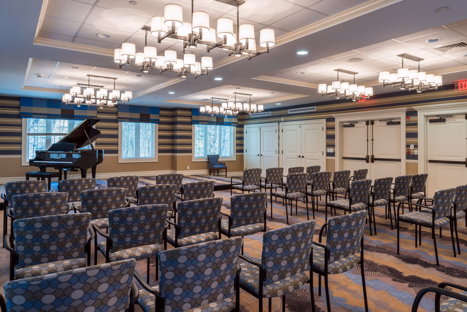 A well-lit room with rows of patterned chairs facing a small stage area with a grand piano. The room has large windows with blue and beige striped valances, multiple modern chandeliers, and beige walls with horizontal striped wallpaper. There are several closed double doors along one wall.