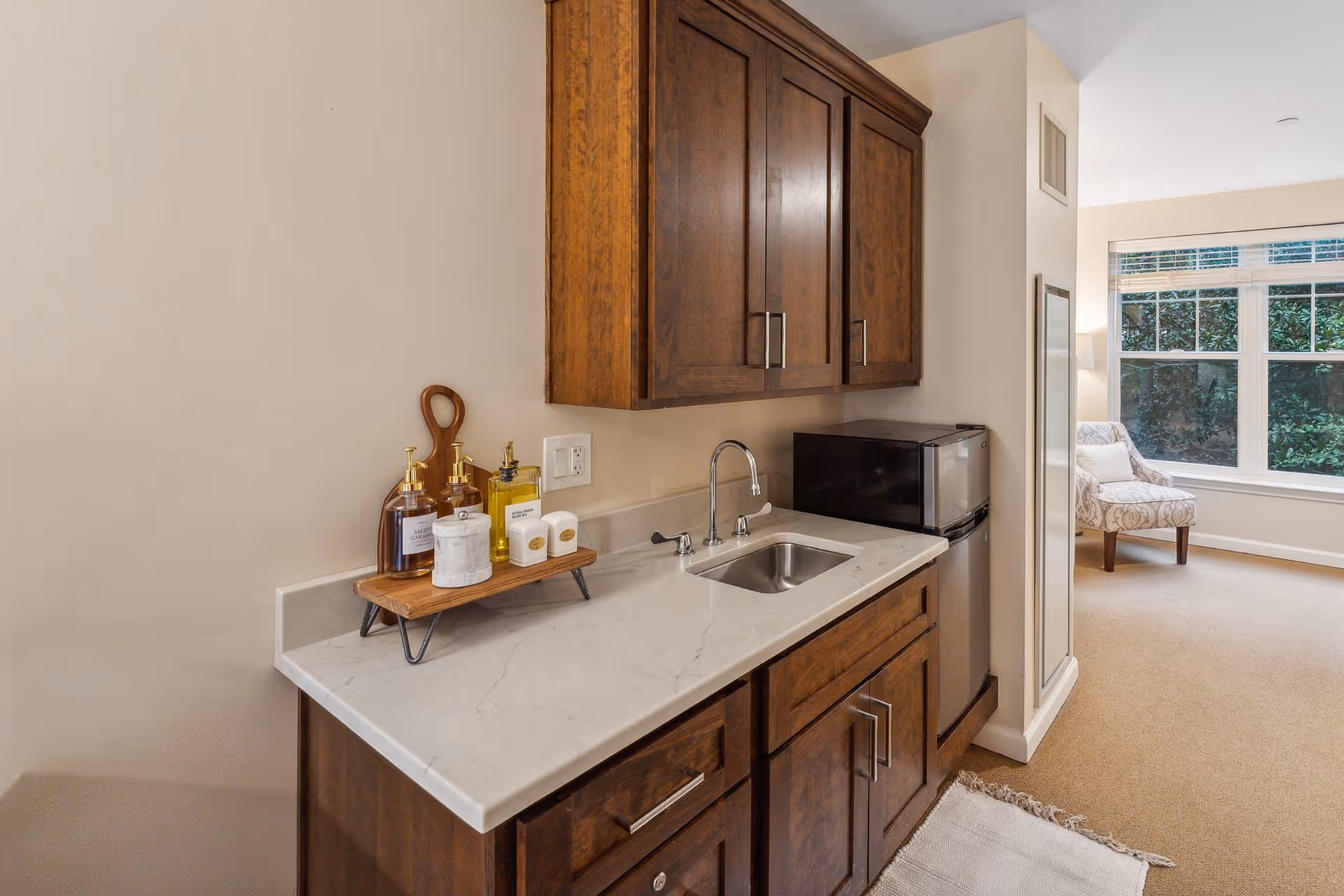 A small kitchenette area with dark wooden cabinets, a white marble countertop, a stainless steel sink, and a mini refrigerator. On the countertop, there are soap dispensers and other small decorative items on a wooden tray. In the background, there is a carpeted room with a patterned armchair near a large window with white blinds.