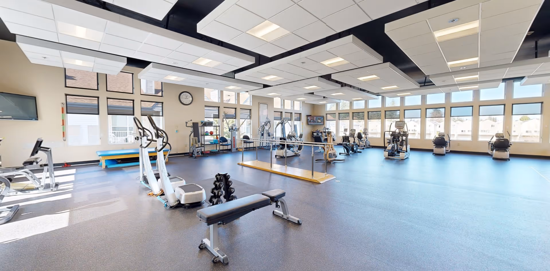A spacious fitness room with large windows letting in natural light. The room contains various exercise equipment including stationary bikes, elliptical machines, a weight bench, dumbbells, and parallel bars for balance exercises. The ceiling has white acoustic panels and the floor is covered with a blue rubberized material.