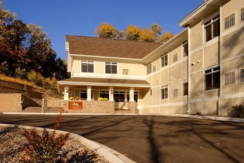 Exterior view of a multi-story assisted living facility building with beige siding and multiple windows. The entrance area has a covered porch with columns and a sign that reads 'Comforts of Home Advanced Assisted Living'. Trees with autumn foliage are visible in the background under a clear blue sky.