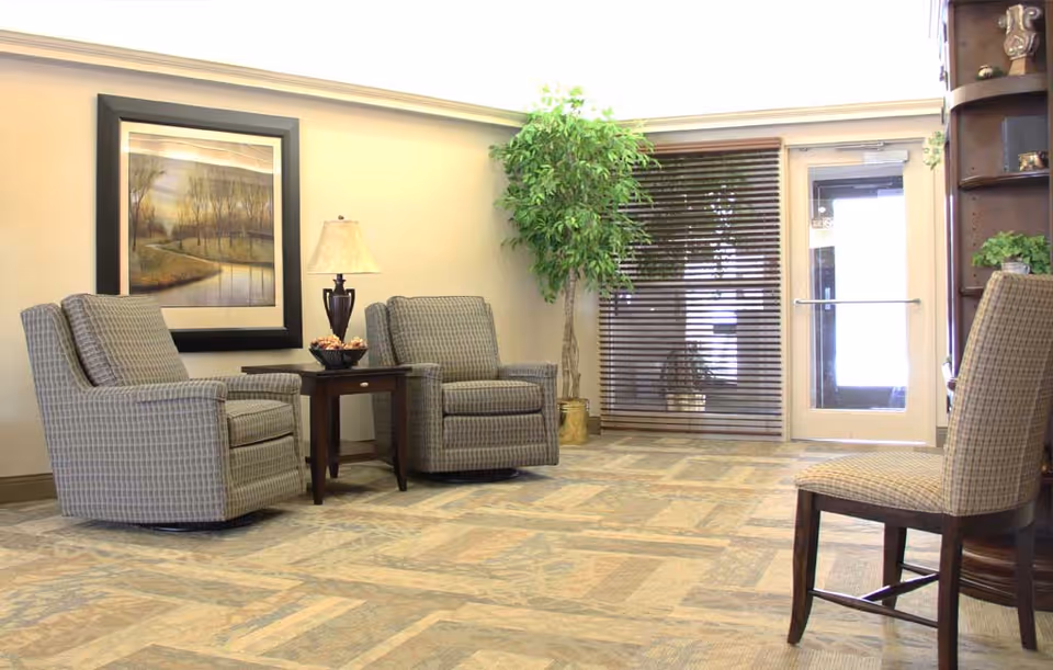 A cozy sitting area in a retirement community featuring two patterned armchairs with a small wooden table and lamp between them, a large framed landscape painting on the wall, a tall potted plant near a glass door with blinds, and a wooden chair partially visible on the right side.
