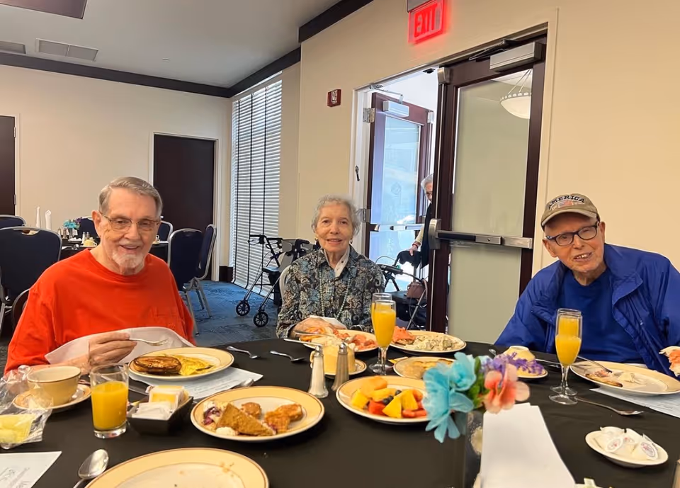 Three elderly people sitting at a dining table with plates of food and glasses of orange juice in front of them inside a dining room. There is a walker near the window and an exit door in the background.