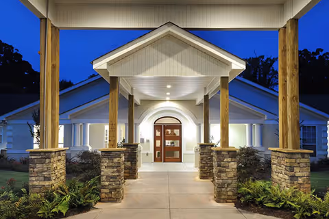 Well-lit covered entrance with wooden posts on stone bases leading to the front doors of a single-story building at dusk.