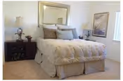 Carpeted bedroom with a neatly made bed topped with multiple pillows, flanked by two bedside tables with lamps, a mirror above the headboard, and framed art on the wall.