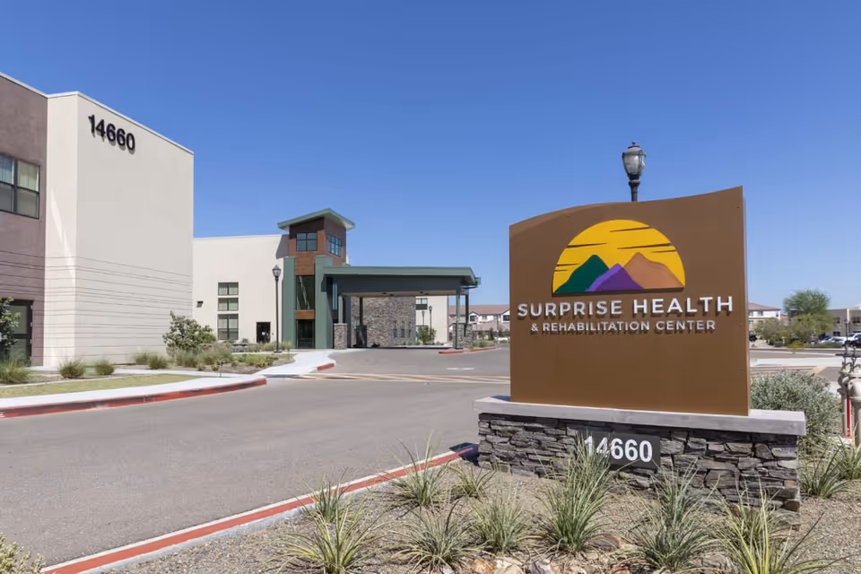 Exterior view of Surprise Health & Rehabilitation Center building with a large sign in the foreground displaying the facility name and logo. The building is modern with beige and brown tones, and the address number 14660 is visible on both the building and the sign. The sky is clear and blue.