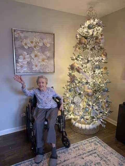 An elderly woman sitting in a wheelchair waving her hand next to a decorated and lit Christmas tree in a cozy room with a floral painting on the wall and a patterned rug on the floor.