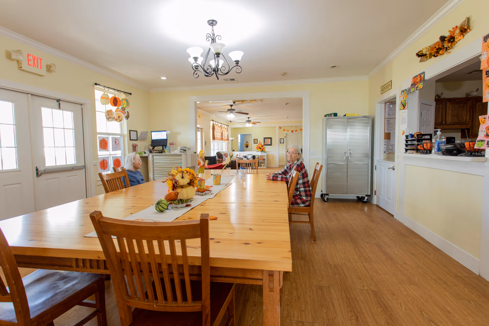 A bright dining room in Mercy House Temple with a long wooden table decorated with autumn-themed centerpieces. Two elderly women are seated at the table. The room has light yellow walls, wooden flooring, a chandelier, and large windows letting in natural light. There are decorations on the walls and a serving window to the right.