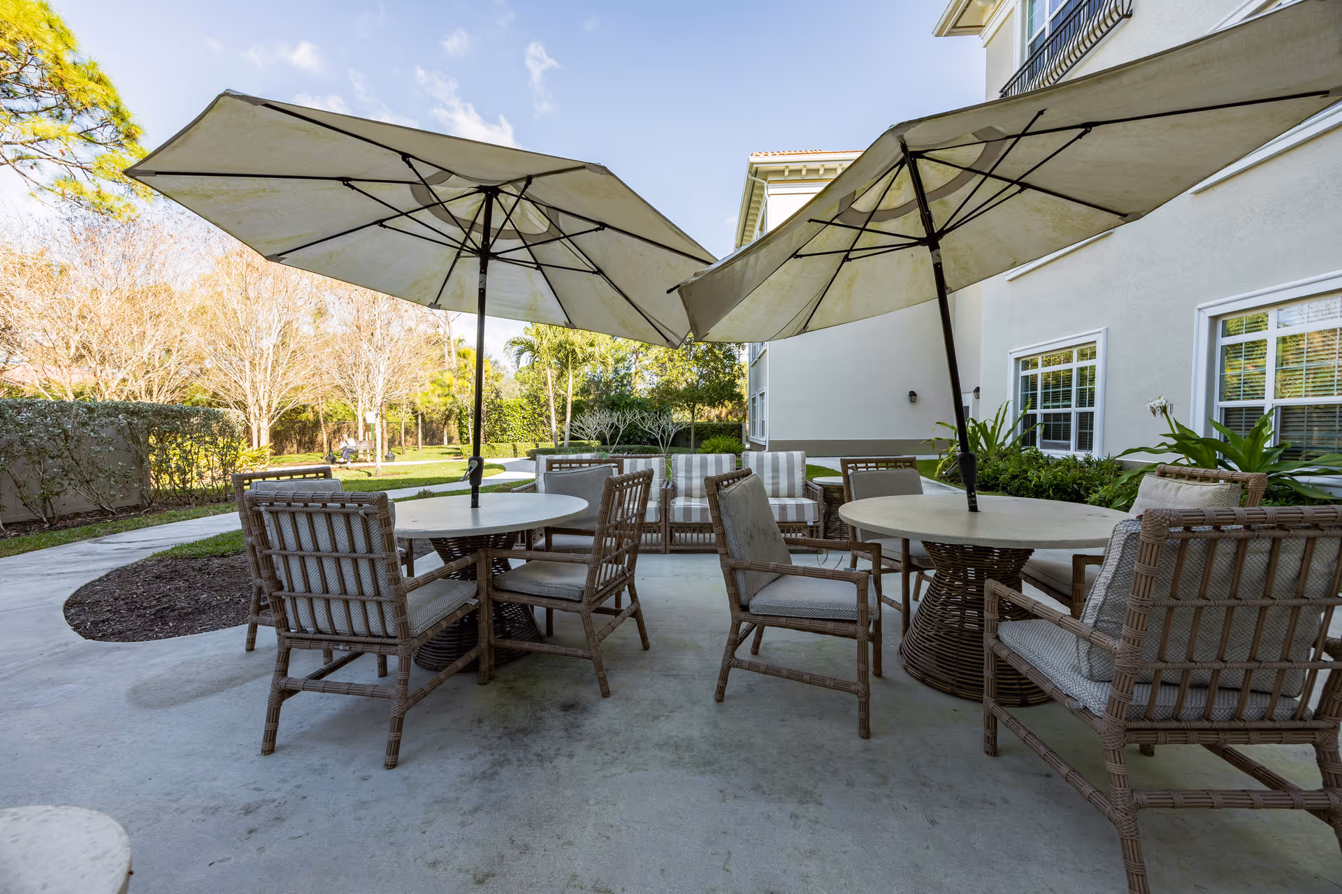 Patio with round tables, wicker chairs, and large umbrellas next to a building and landscaped yard.
