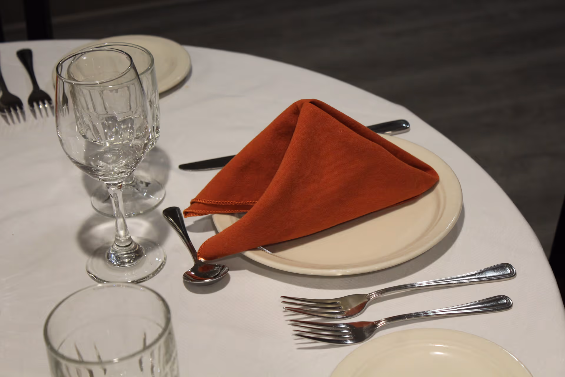 A close-up view of a dining table set with a white tablecloth, a beige plate with a folded rust-colored cloth napkin, silverware including forks, a spoon, and a knife, and multiple clear drinking glasses.