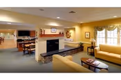 A cozy living room area in a senior living facility with beige sofas, a coffee table, a fireplace with a stone base, and a TV mounted on the wall. The room is well-lit with natural light coming through a large window with curtains. There are decorative items on the mantle and framed pictures on the walls.