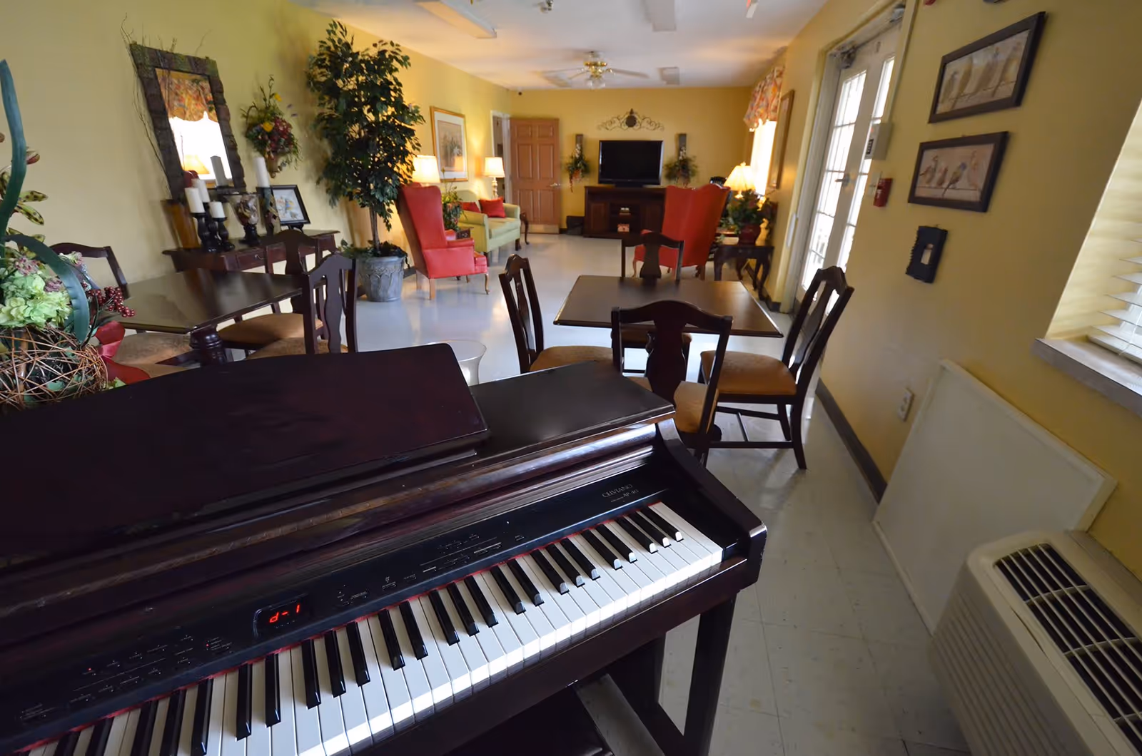 Interior view of a common area in a senior living facility with a digital piano in the foreground, several wooden tables and chairs, red armchairs, a green sofa, decorative plants, framed pictures on yellow walls, and a television on a stand at the far end.