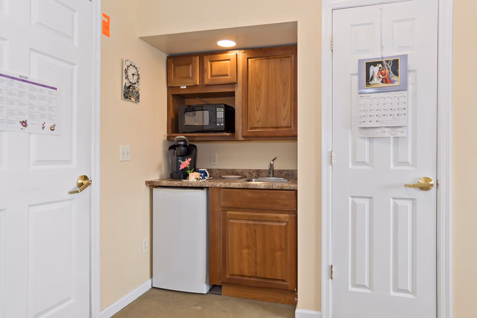 Small kitchenette area with wooden cabinets, a countertop with a coffee maker, a small refrigerator, a microwave, and a sink. Two white doors flank the kitchenette, each with a calendar hanging on it. The walls are painted beige.
