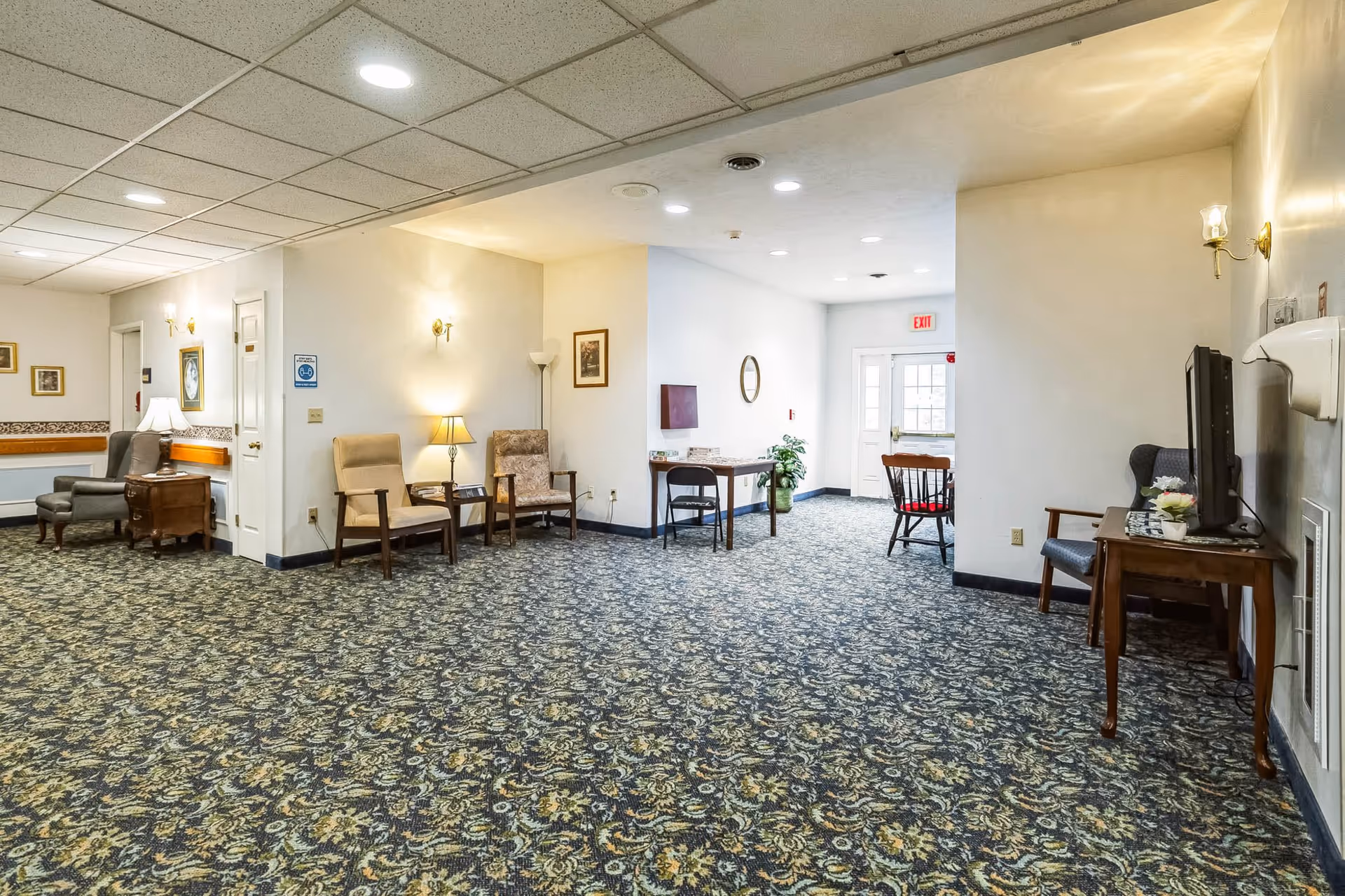 A spacious interior common area with patterned carpet, several armchairs, a small table with a television, wall sconces, and a door leading outside. The area is well-lit with ceiling lights and lamps, and there are framed pictures on the walls.
