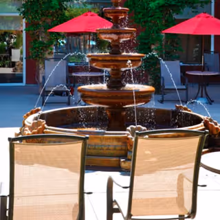 Outdoor courtyard area with a multi-tiered water fountain in the center, surrounded by patio chairs and tables with red umbrellas. Greenery and building windows are visible in the background.