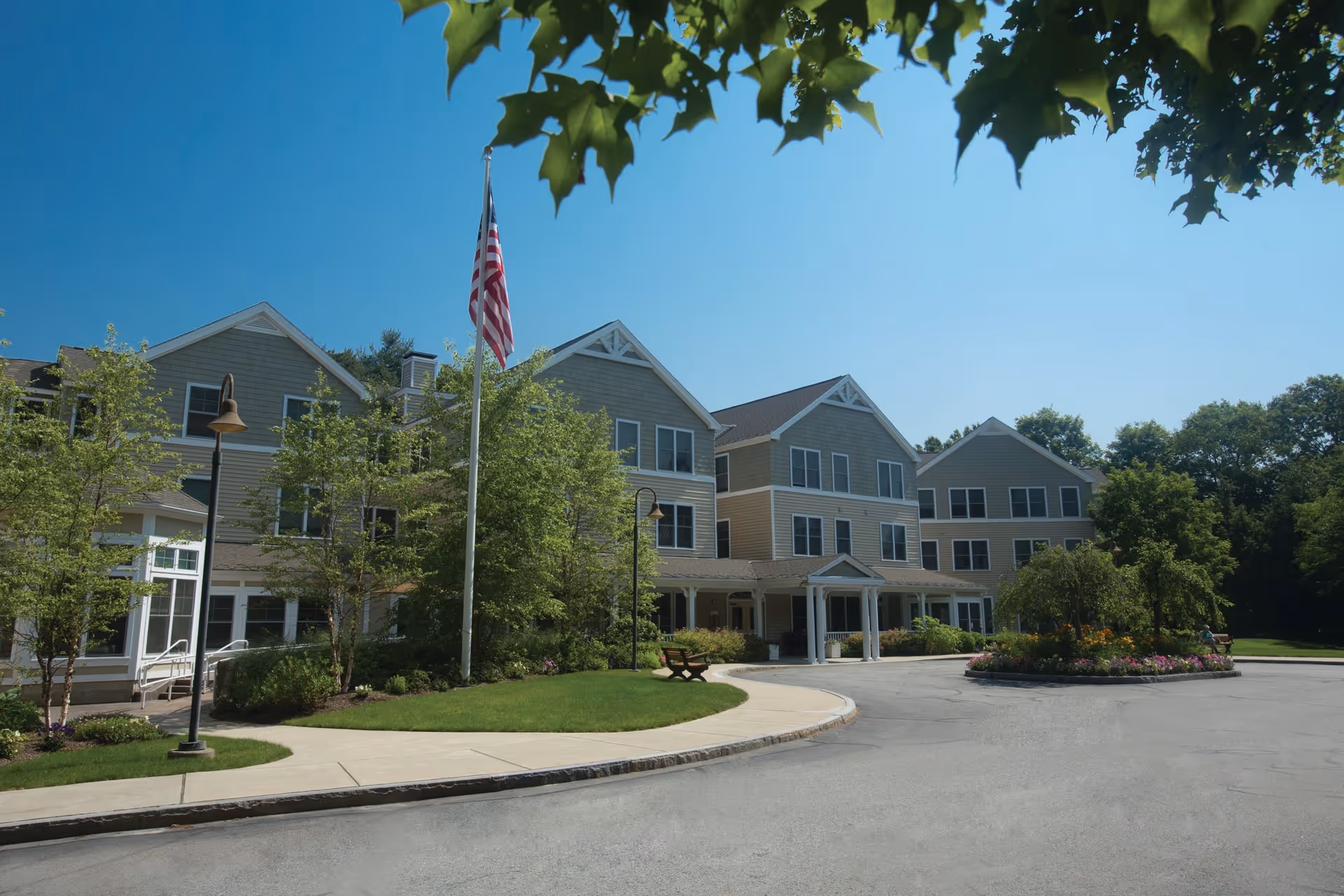 Exterior view of a multi-story senior living facility building with beige siding and white trim, surrounded by trees and landscaping. An American flag is flying on a flagpole near the entrance, which has a covered drop-off area. The sky is clear and blue.