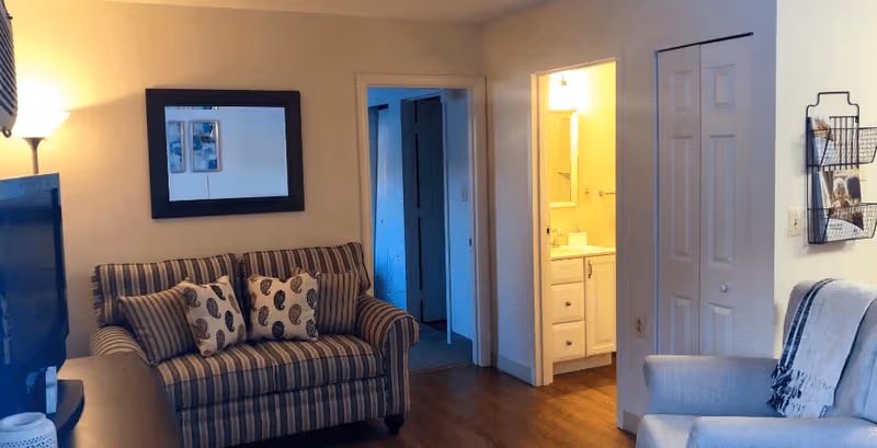 A cozy living room area with a striped loveseat adorned with patterned pillows, a floor lamp, a wall mirror, and a glimpse into a bathroom with a sink and mirror. There is also a white armchair with a throw blanket and a wall-mounted magazine rack.