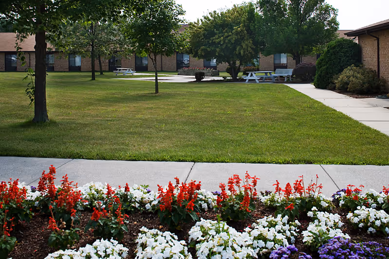 A well-maintained outdoor garden area at Care Haven Center featuring a flower bed with red, white, and purple flowers in the foreground, a green lawn with several trees, white picnic tables, and a brick building with windows in the background.
