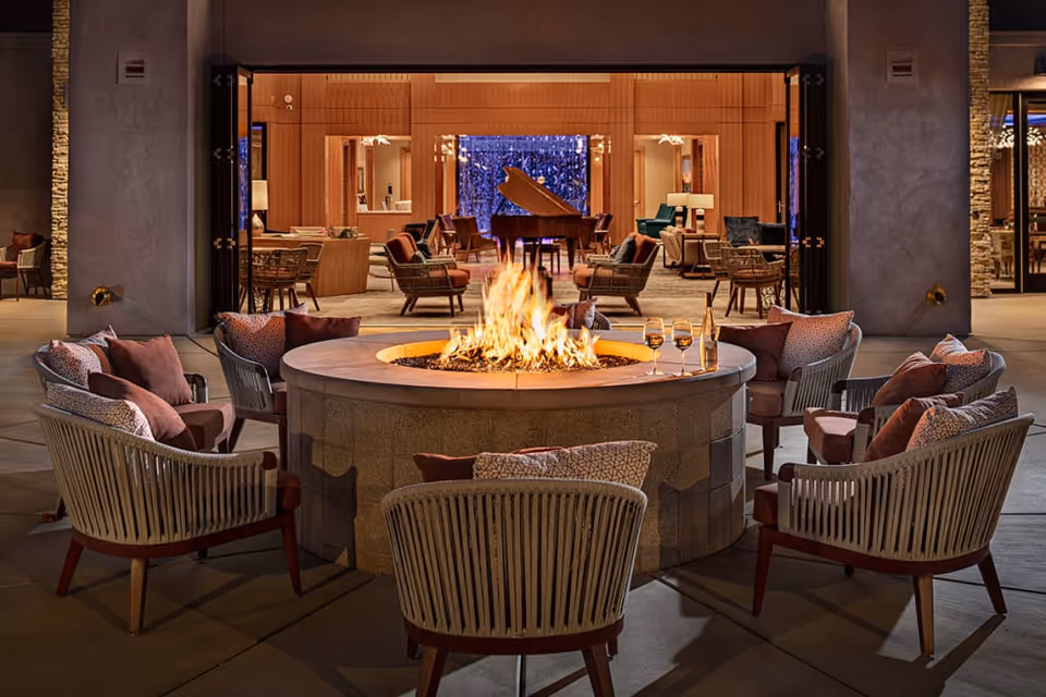 Outdoor seating area at night with cushioned chairs arranged around a large circular fire pit. In the background, an open indoor lounge area is visible with more chairs, tables, and a grand piano under warm lighting.