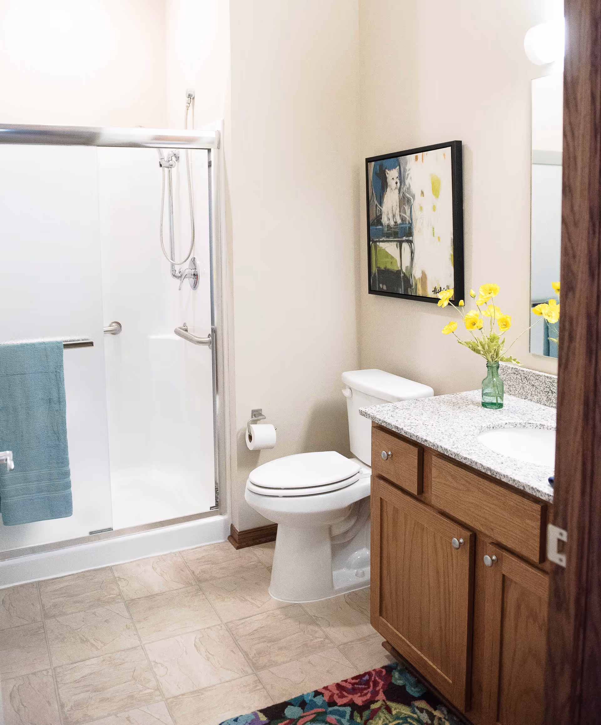 A clean bathroom with a glass-enclosed shower, a white toilet, a wooden vanity with a granite countertop, a mirror, a vase with yellow flowers, a colorful rug on the floor, and a framed picture of a dog on the wall.