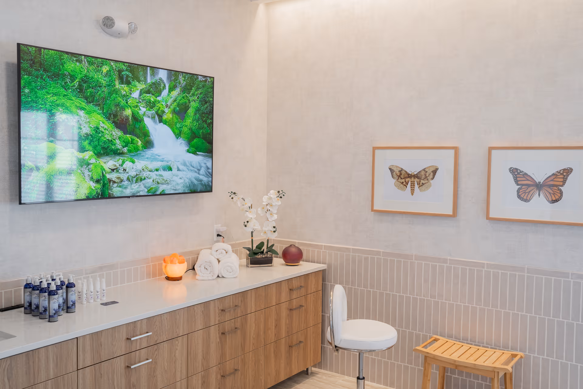 Bright, spa-like interior with a wall-mounted TV showing a waterfall, wooden cabinets topped with rolled towels, orchids and toiletries, a stool and framed butterfly prints on the wall.