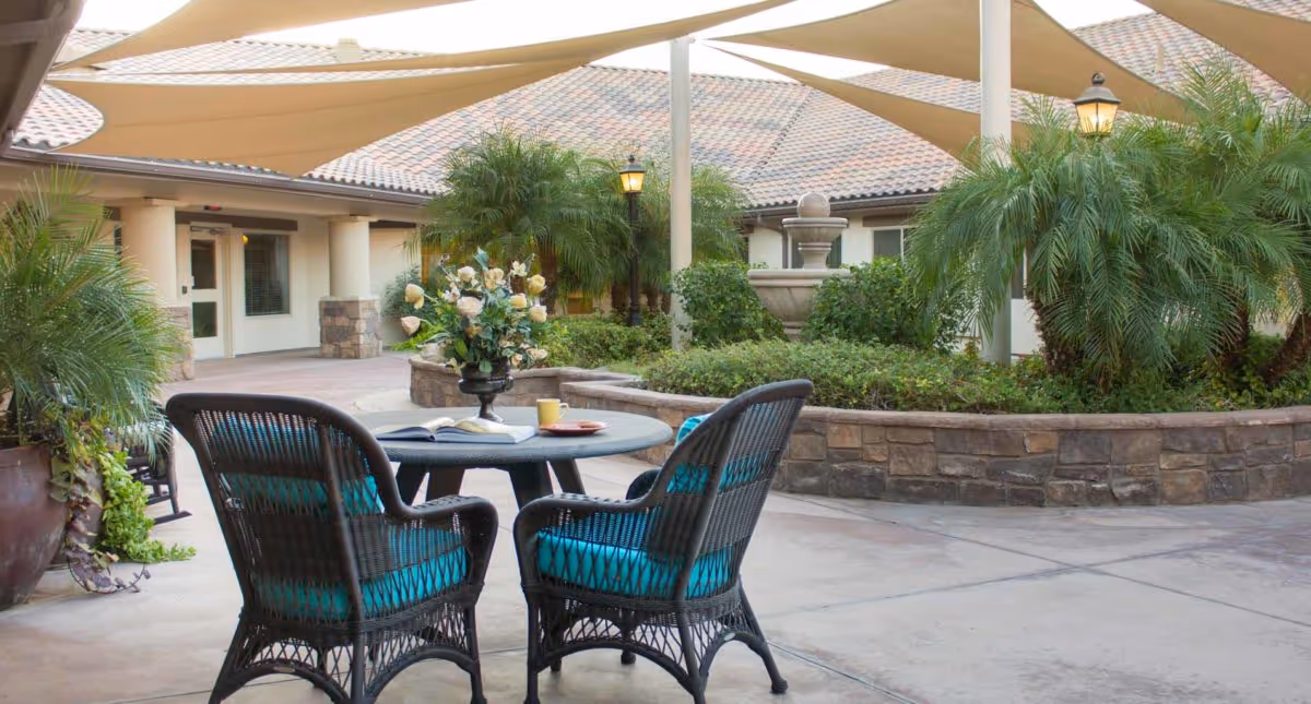 Outdoor patio area with a round table and two wicker chairs with blue cushions. On the table, there is a vase with flowers, an open book, a yellow mug, and a red plate. The patio is surrounded by greenery, palm plants, and a stone wall with a fountain in the background. Shade sails are overhead, and the building with tiled roofs is visible.
