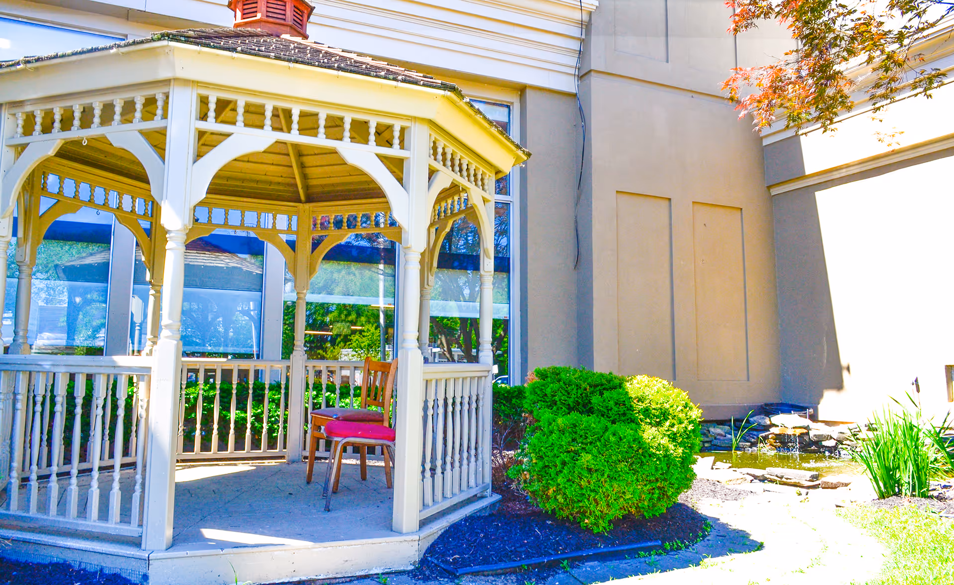 White wooden gazebo with a chair and red cushion next to a building and landscaped shrubs.