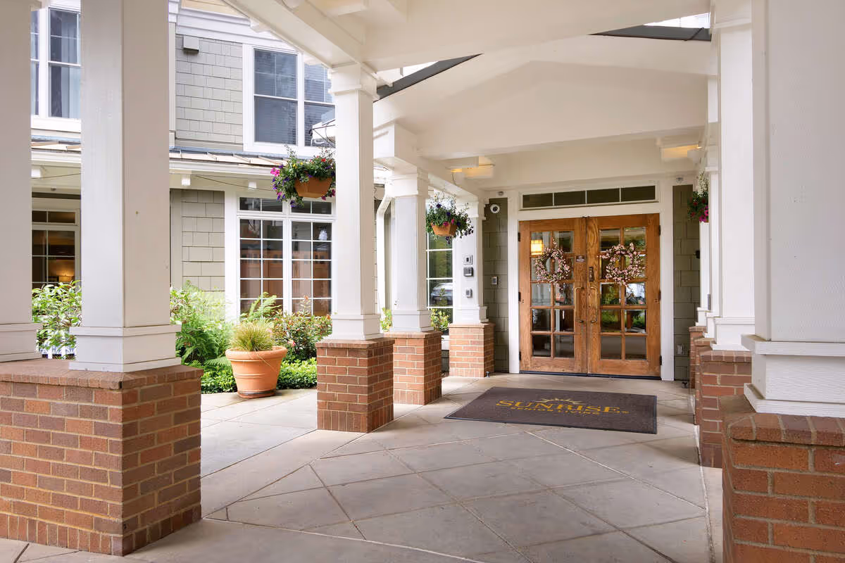 Covered front entrance of a senior living facility with brick columns, hanging plants, a welcome mat, and wooden double doors.