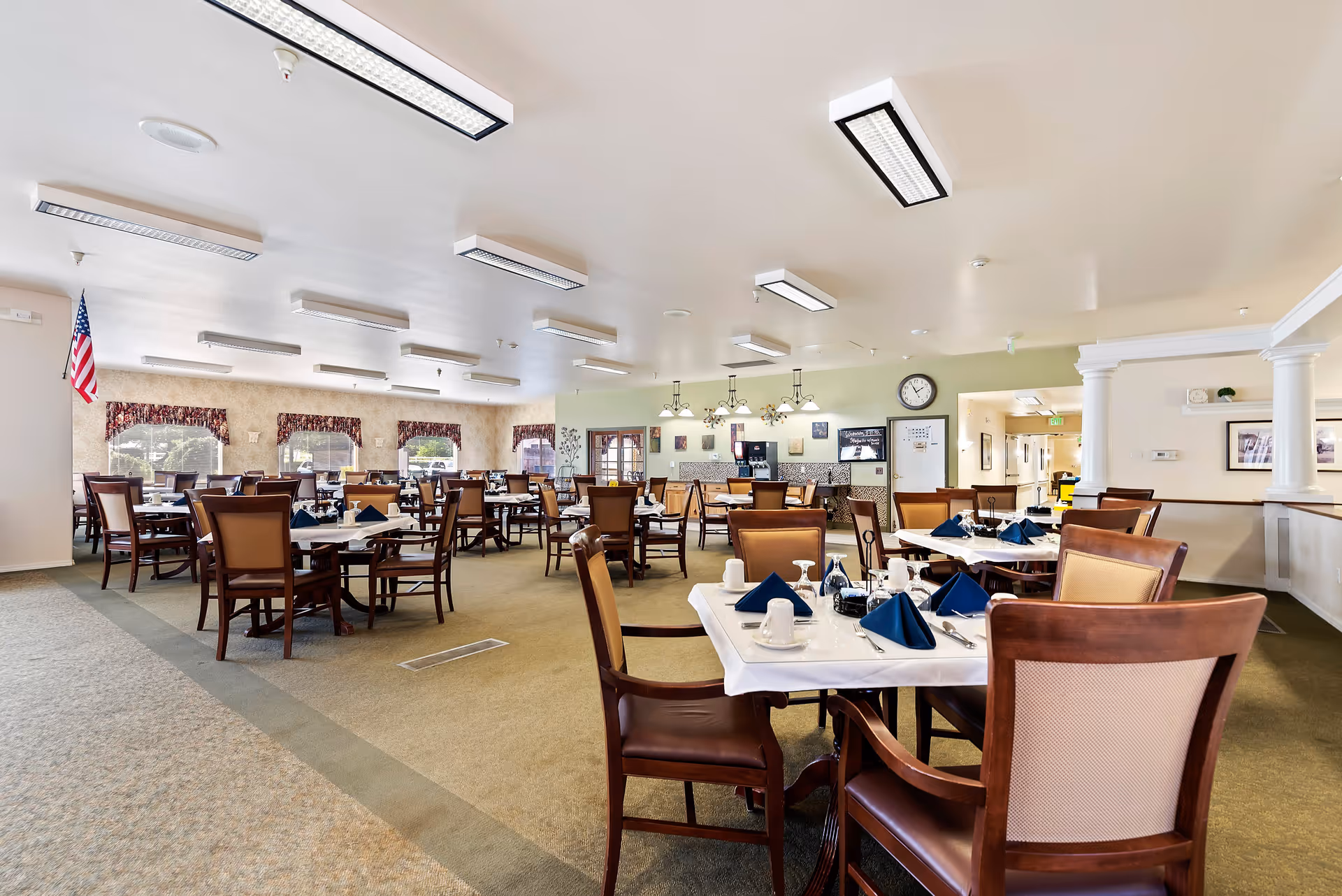 A spacious dining room in an assisted living facility with multiple tables set with white tablecloths, blue folded napkins, cups, and glasses. The room has beige carpeting, several windows with valances, and overhead fluorescent lighting. There is an American flag in the corner and a clock on the wall above a counter area.