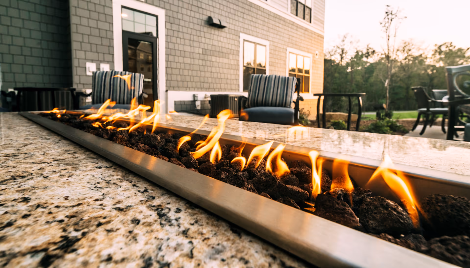 Outdoor patio area with a long rectangular fire pit built into a granite countertop, surrounded by cushioned chairs and tables, with a building and trees in the background during sunset.