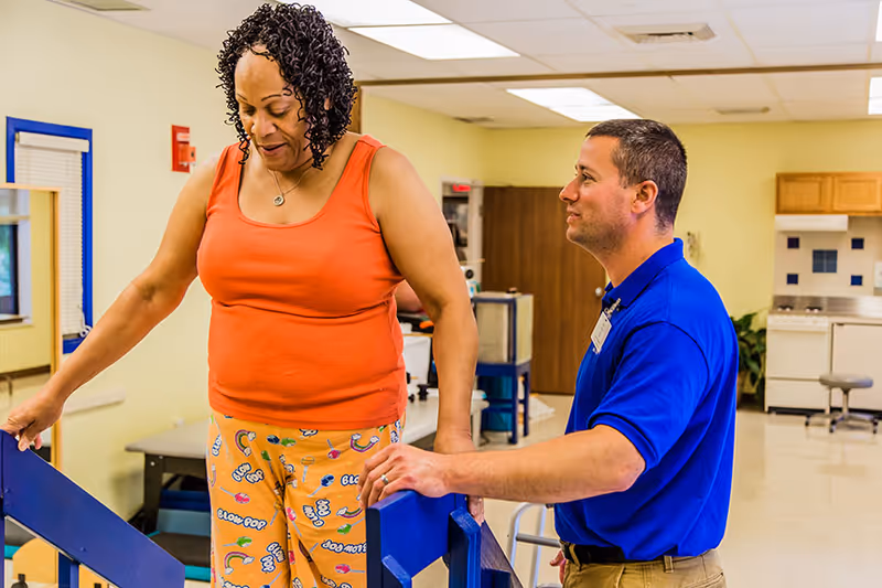 A woman in an orange tank top and colorful pajama pants is walking on parallel bars for physical therapy while a male therapist in a blue shirt assists her in a rehabilitation center room with medical and therapy equipment.