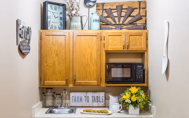 Small kitchenette with oak cabinets, a sink, microwave, decorative signs and a vase of yellow flowers on the counter.