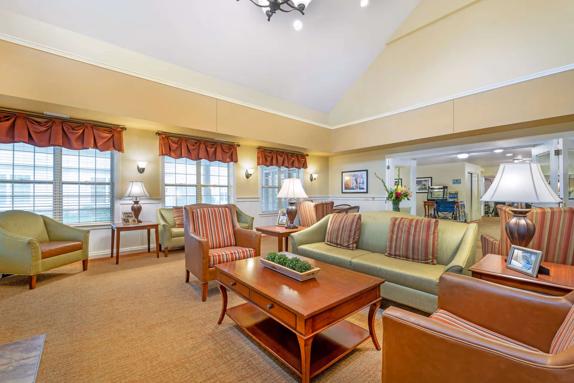 A bright and spacious living room area with beige walls and carpeted floor. The room features a green sofa with striped cushions, two striped armchairs with brown leather sides, and two green armchairs. There are wooden coffee and side tables with lamps and framed photos. Large windows with red valances let in natural light. In the background, there is an open doorway leading to another room with a wheelchair visible.