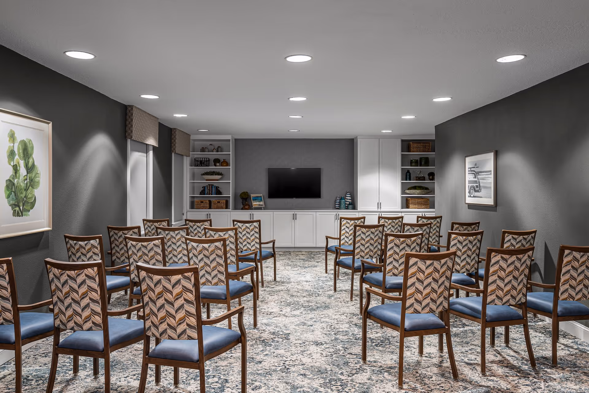 A well-lit meeting or presentation room with multiple wooden chairs arranged in rows facing a wall-mounted flat screen TV. The room has dark gray walls, built-in white cabinets and shelves with decorative items, and framed artwork on the walls. The floor is covered with a patterned carpet.