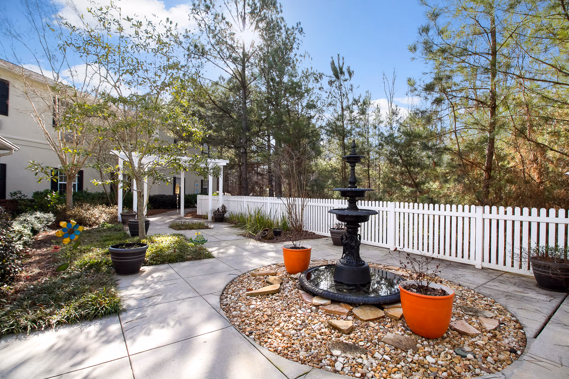 Outdoor garden area with a black tiered fountain surrounded by small rocks and two orange plant pots. There is a white picket fence in the background, trees, and a paved walkway with additional potted plants and greenery. The sky is clear and blue.
