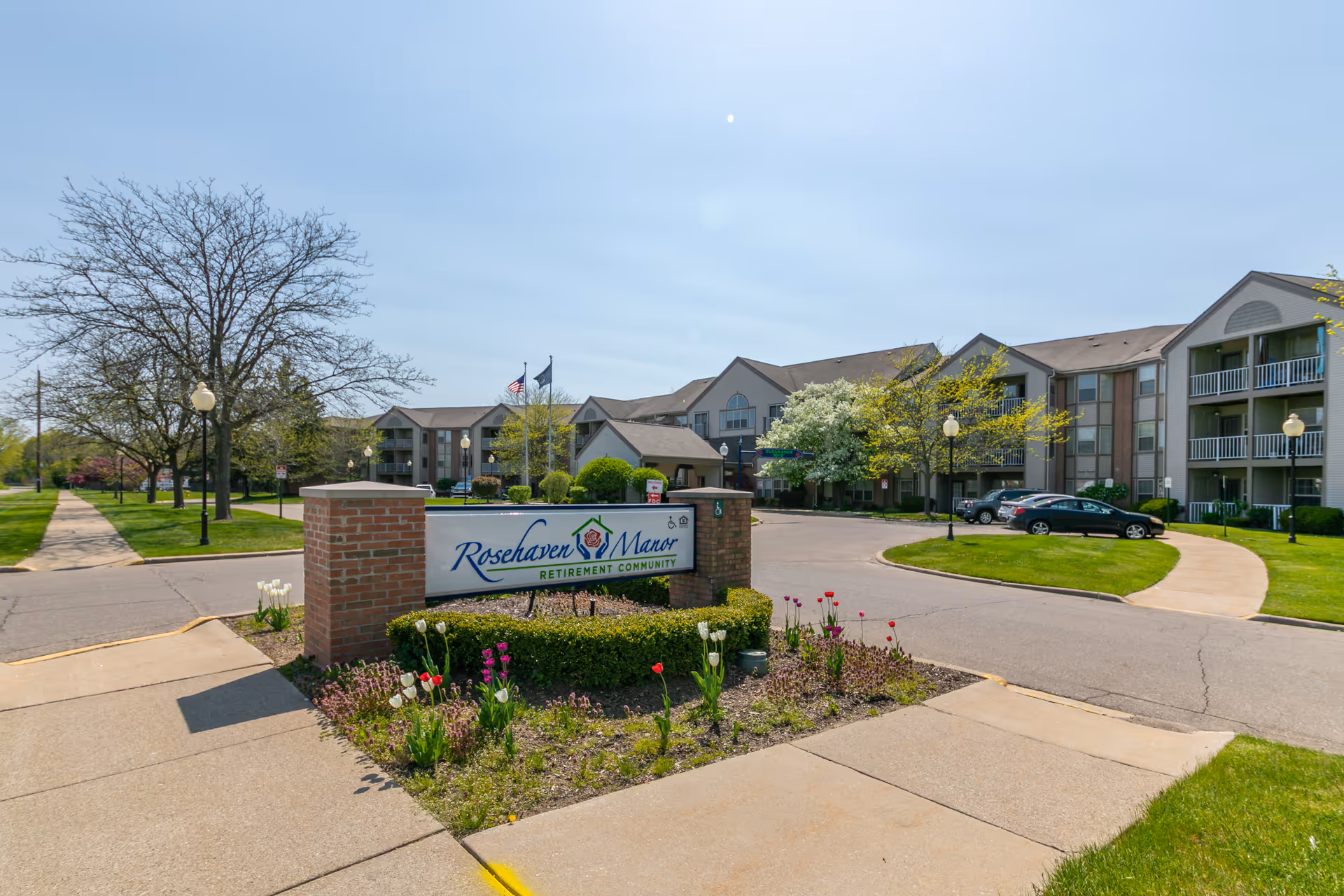 Exterior view of Rosehaven Manor retirement community showing a large building with multiple floors, balconies, and a parking area. The entrance is marked by a brick sign with the community's name, surrounded by flower beds and greenery. Trees and a sidewalk line the street nearby under a clear sky.