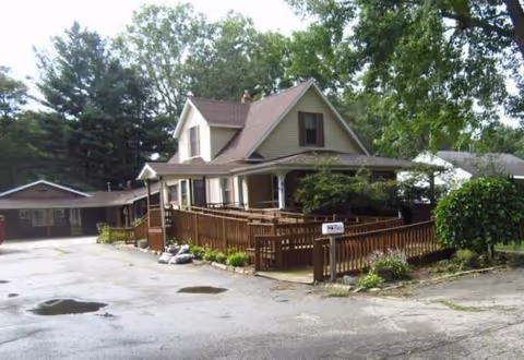Exterior view of a brown, two-story house with a wooden ramp and porch among trees.