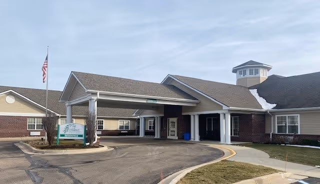 Exterior view of The Willows at East Lansing senior living facility showing the main entrance with a covered driveway, an American flag on a flagpole, and a sign with the facility name. The building has a brick and beige siding exterior with a cupola on the roof. There is a curved driveway and some patches of snow on the roof.