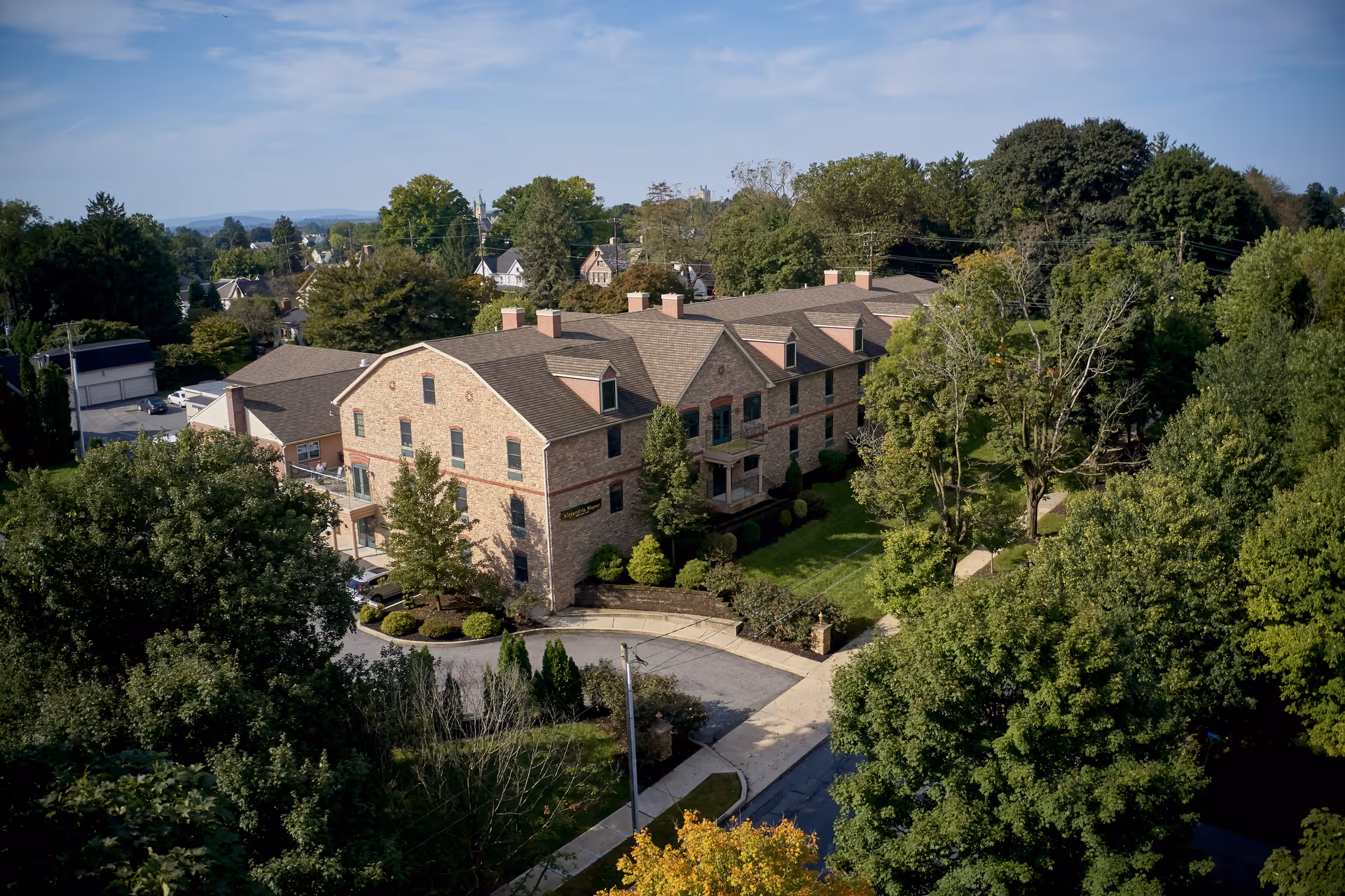 Aerial view of a large brick senior living facility named Alexandria Manor surrounded by trees and greenery, with a driveway and sidewalk leading to the entrance.