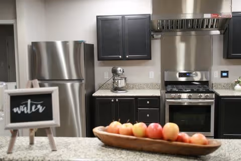 Modern kitchen with stainless steel refrigerator, stove with hood, dark cabinets, granite countertops, a stand mixer, and a wooden bowl filled with apples on the counter. A small chalkboard sign labeled 'water' is also visible on the counter.