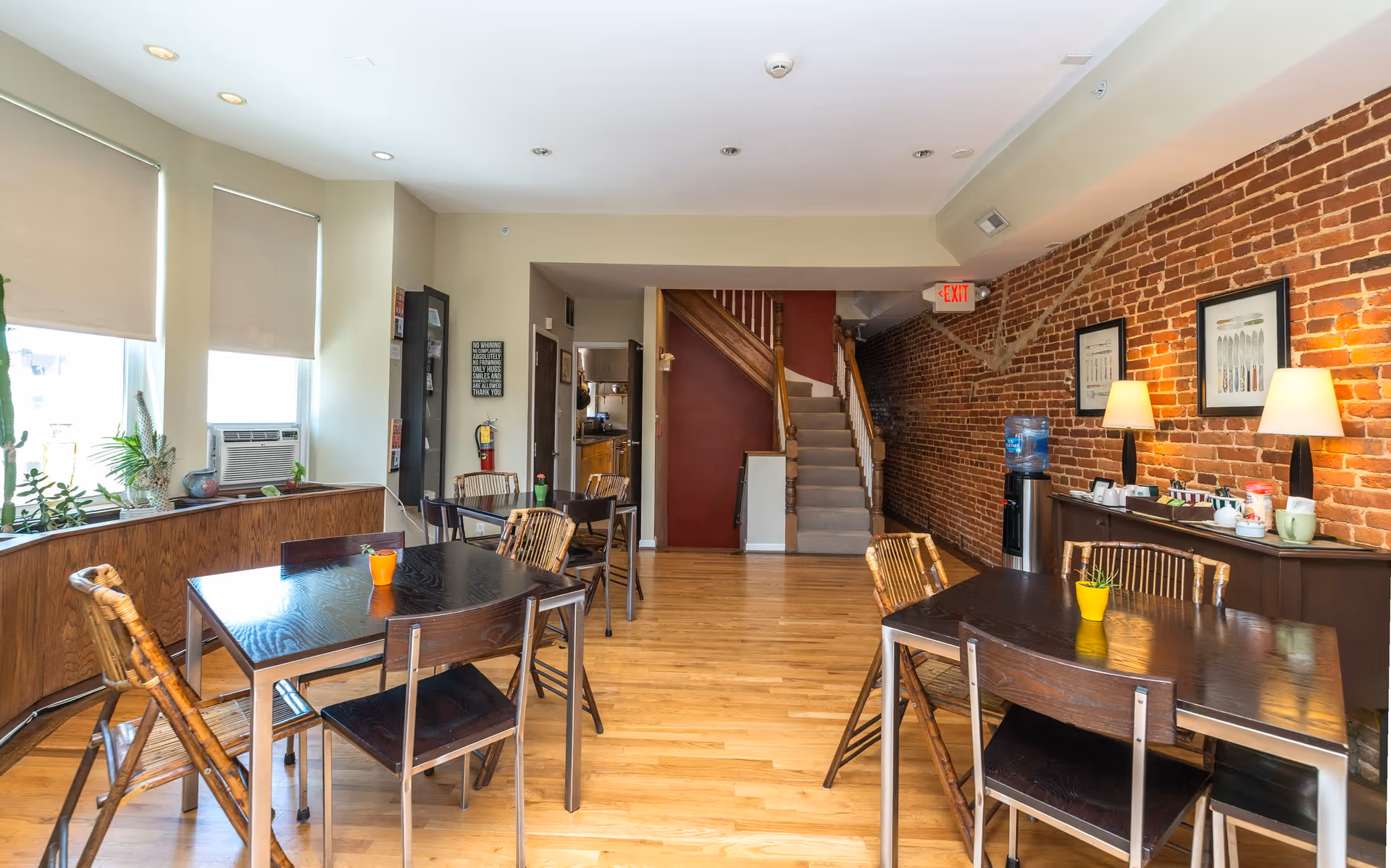 A bright dining area with wooden floors, several dark wood tables and chairs, potted plants on the windowsill, and a brick accent wall with framed artwork and two table lamps. A water cooler and a sideboard with coffee supplies are against the brick wall. A staircase with wooden railing leads to an upper floor, and an exit sign is visible above a hallway.
