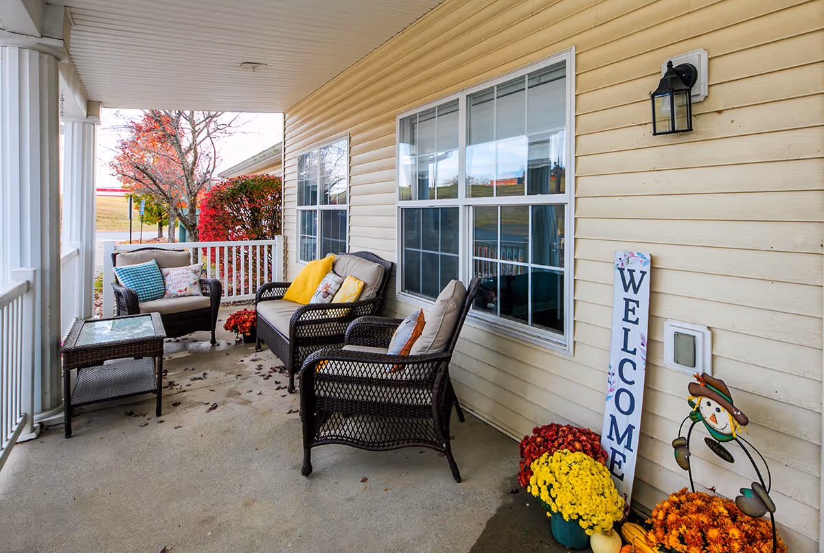 Covered front porch with wicker chairs and cushions, a 'WELCOME' sign, and potted mums.
