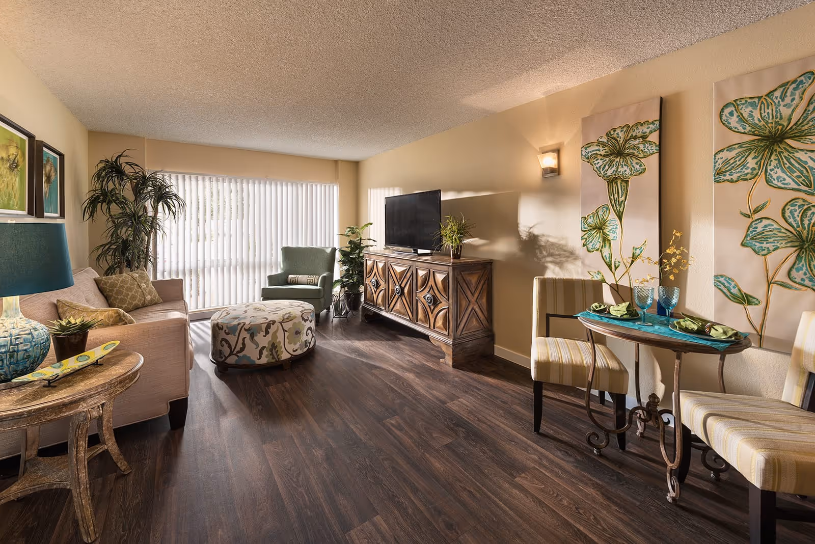 A cozy senior living room with a beige sofa, a patterned ottoman, a green armchair, and a wooden TV cabinet with a flat-screen TV. There is a small round table with two striped chairs set for dining, decorated with blue glasses and green napkins. The room has dark wood flooring, large windows with vertical blinds, and floral artwork on the walls.