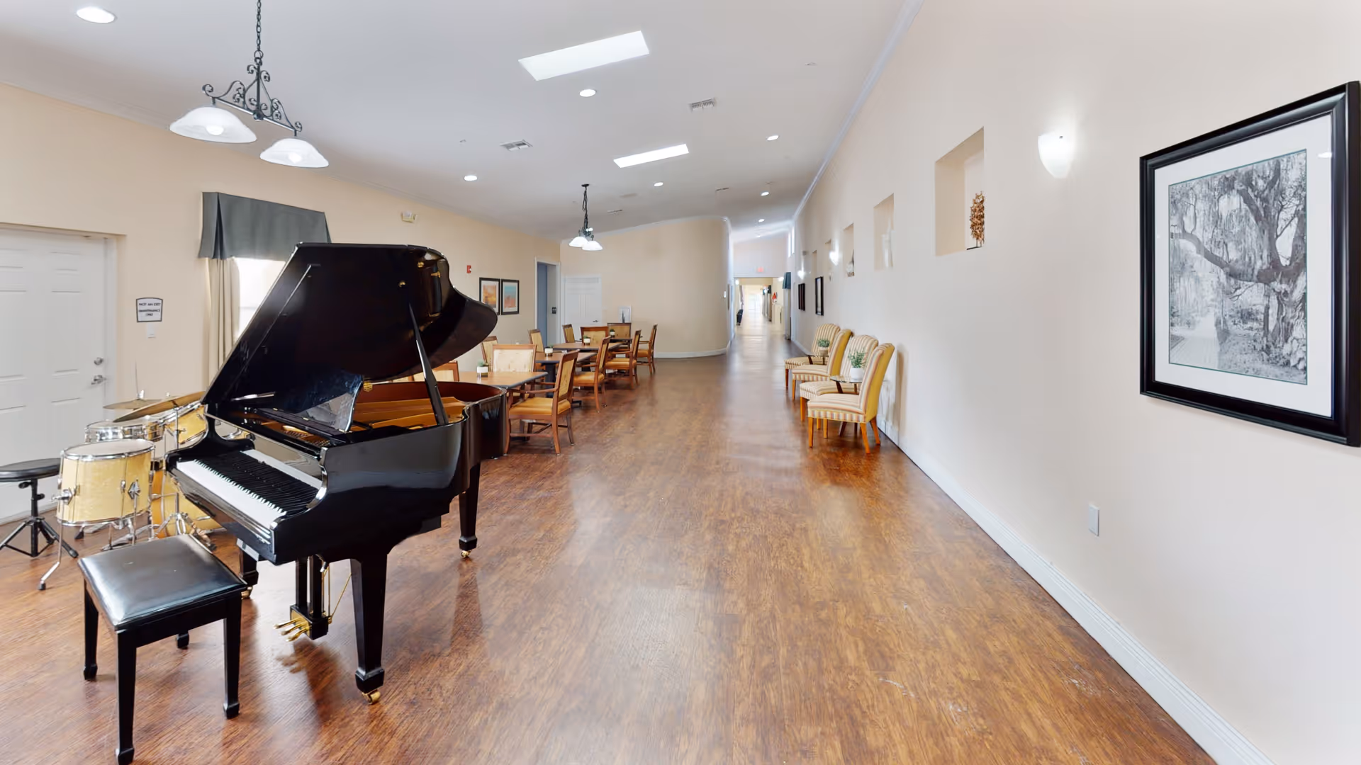 Spacious interior common area with a black grand piano and drum set in the foreground, dining tables and chairs down the center, and framed artwork on the right wall.