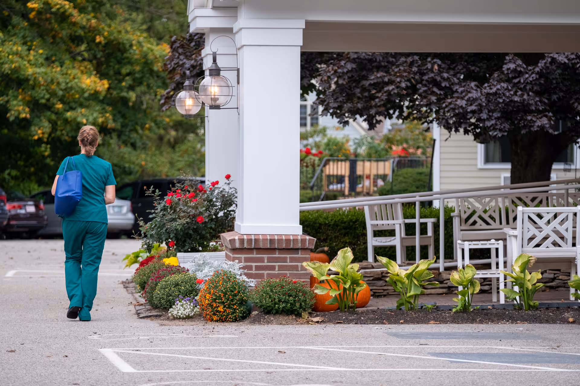 A person wearing green scrubs and carrying a blue bag walks away from the camera along a paved area next to a building entrance with white columns and brick bases. There are colorful flowers and pumpkins planted along the edge of the walkway, and white outdoor chairs and tables are visible under a tree near the building. Several parked cars and green trees are in the background.