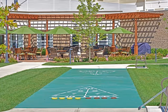 Outdoor courtyard with a pergola, patio seating and green umbrellas, and a shuffleboard court in the foreground.