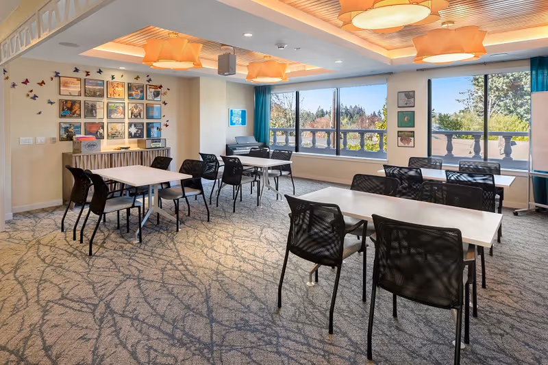 A bright and spacious room with several tables and black chairs arranged for group seating. The room has large windows with a view of trees and greenery outside. The walls are decorated with framed pictures and colorful butterfly decorations. The ceiling features modern light fixtures, and the carpet has a subtle abstract pattern.