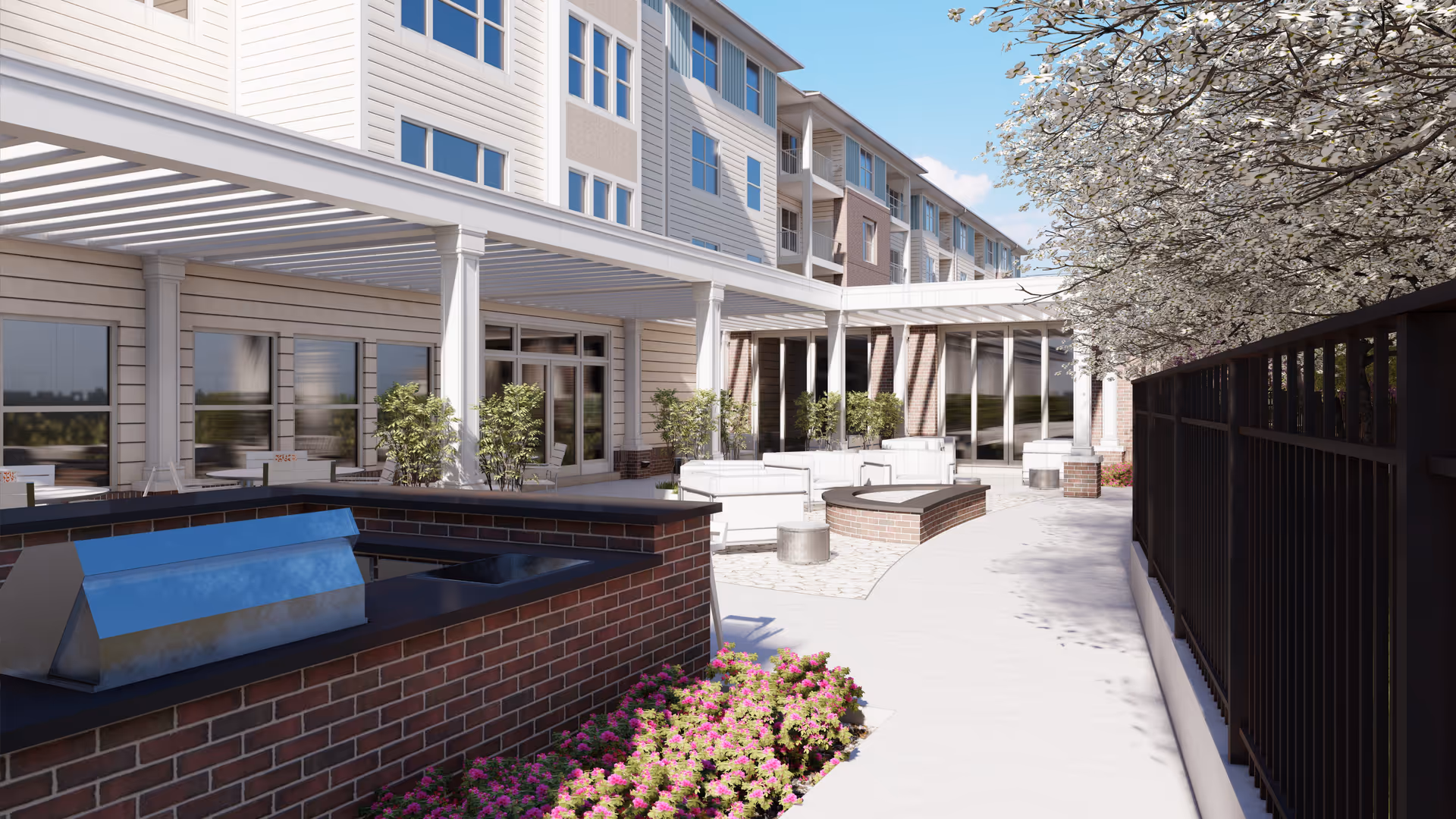 Outdoor patio area of a senior living facility with a built-in brick grill, white cushioned seating, potted plants, and flowering trees under a clear blue sky. The building has multiple floors with large windows and a covered walkway supported by white columns.